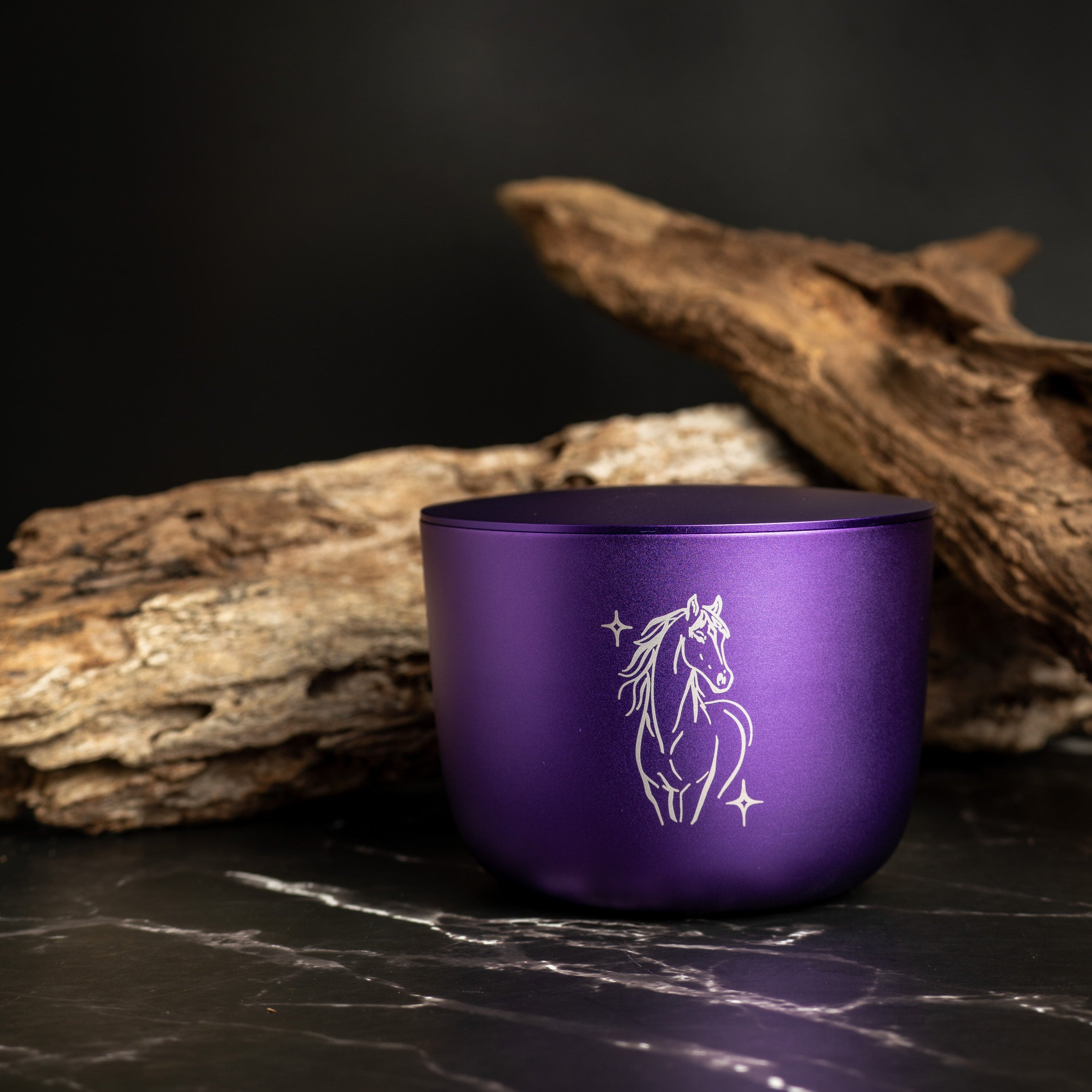 Purple soy candle with a horse design on a dark surface with wooden logs.