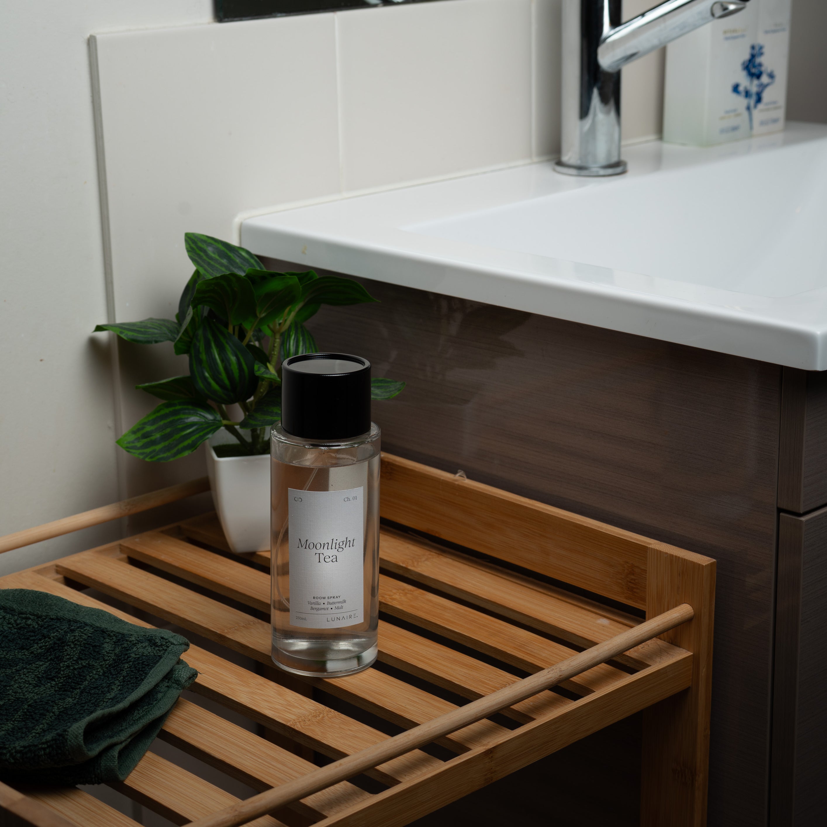 A clear glass bottle with a black cap and white label sits on a wooden slatted shelf beside a white sink, with a folded dark green towel and a small potted plant nearby. Room Refresher Spray