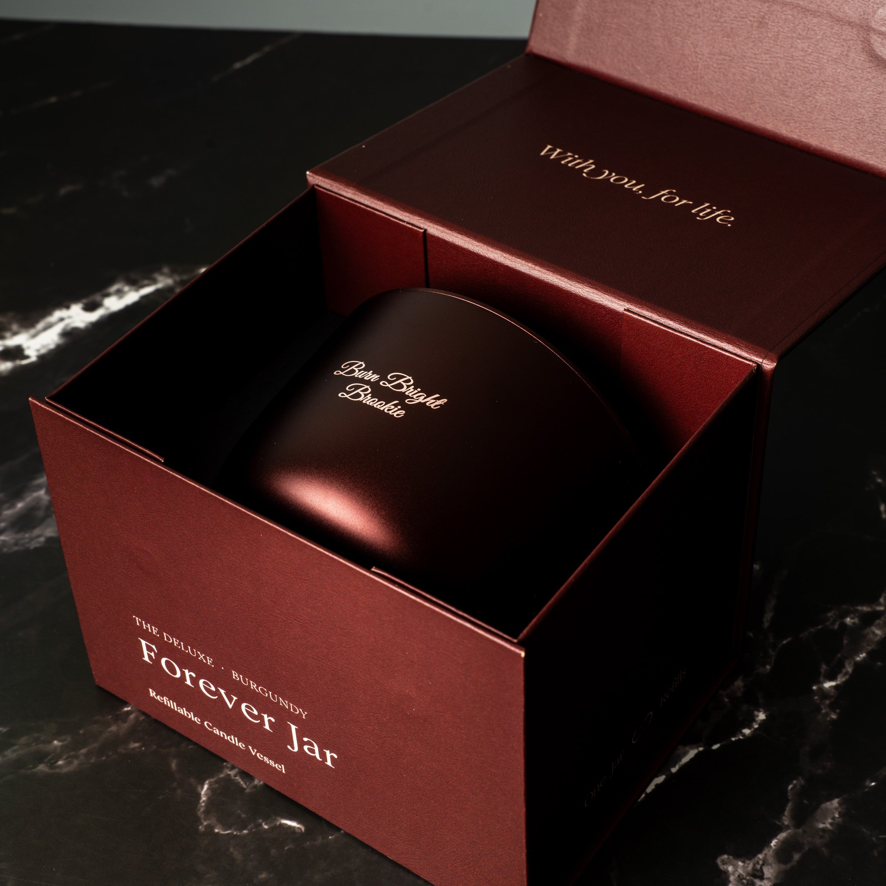 burgundy-colored candle jar with 'Forever Jar' branding inside an open red box on a dark marble surface.