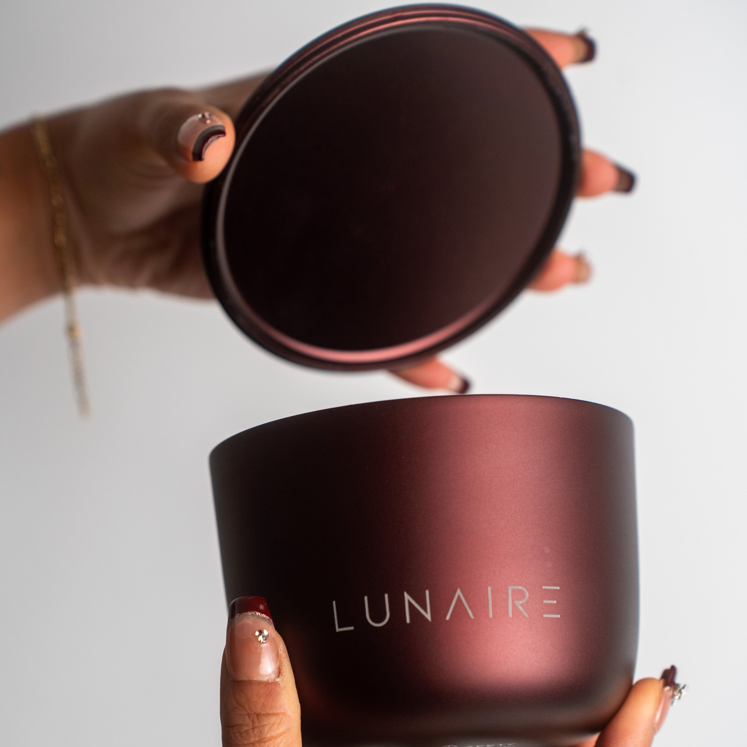 Person holding a dark red candle jar with 'LUNAIRE' branding against a light background