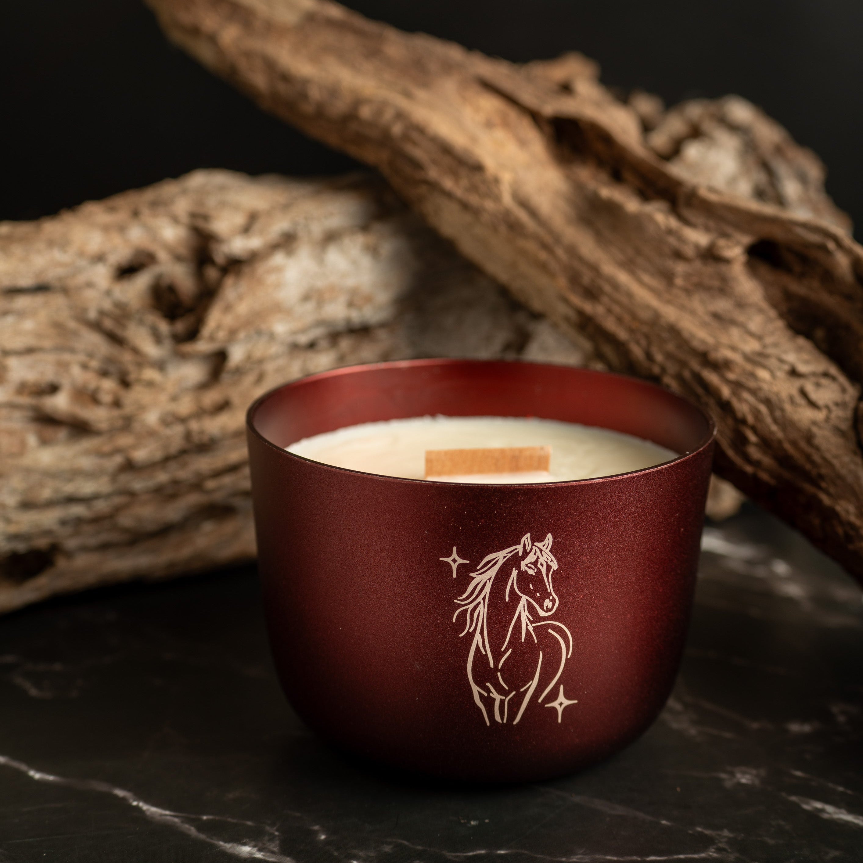 Burgundy aluminium candle holder with a horse design on a dark surface with driftwood.