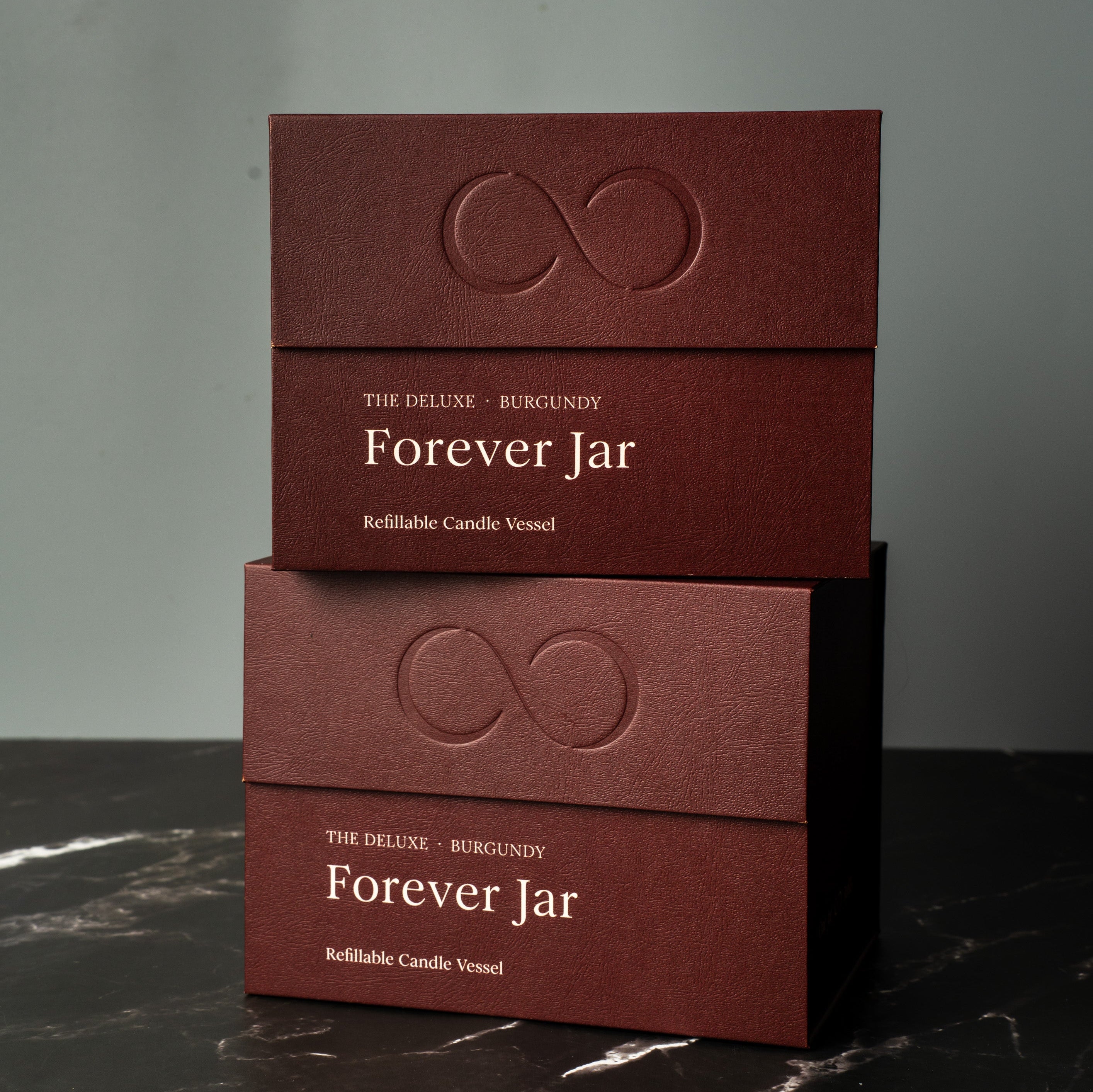 Two burgundy boxes labeled 'Forever Jar' on a dark marble surface with a gray background. Premium Gift Box candles