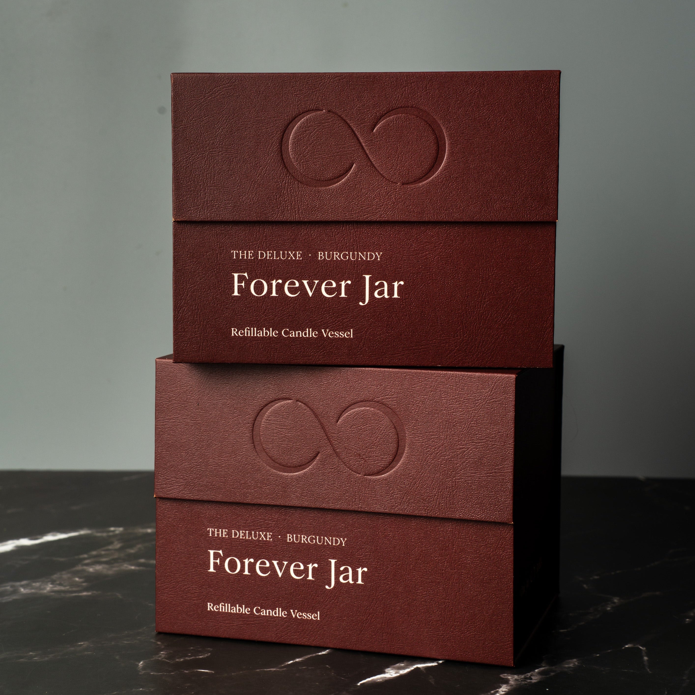 Two burgundy boxes labeled 'Forever Jar' on a dark marble surface with a gray background. Premium Gift Box candles