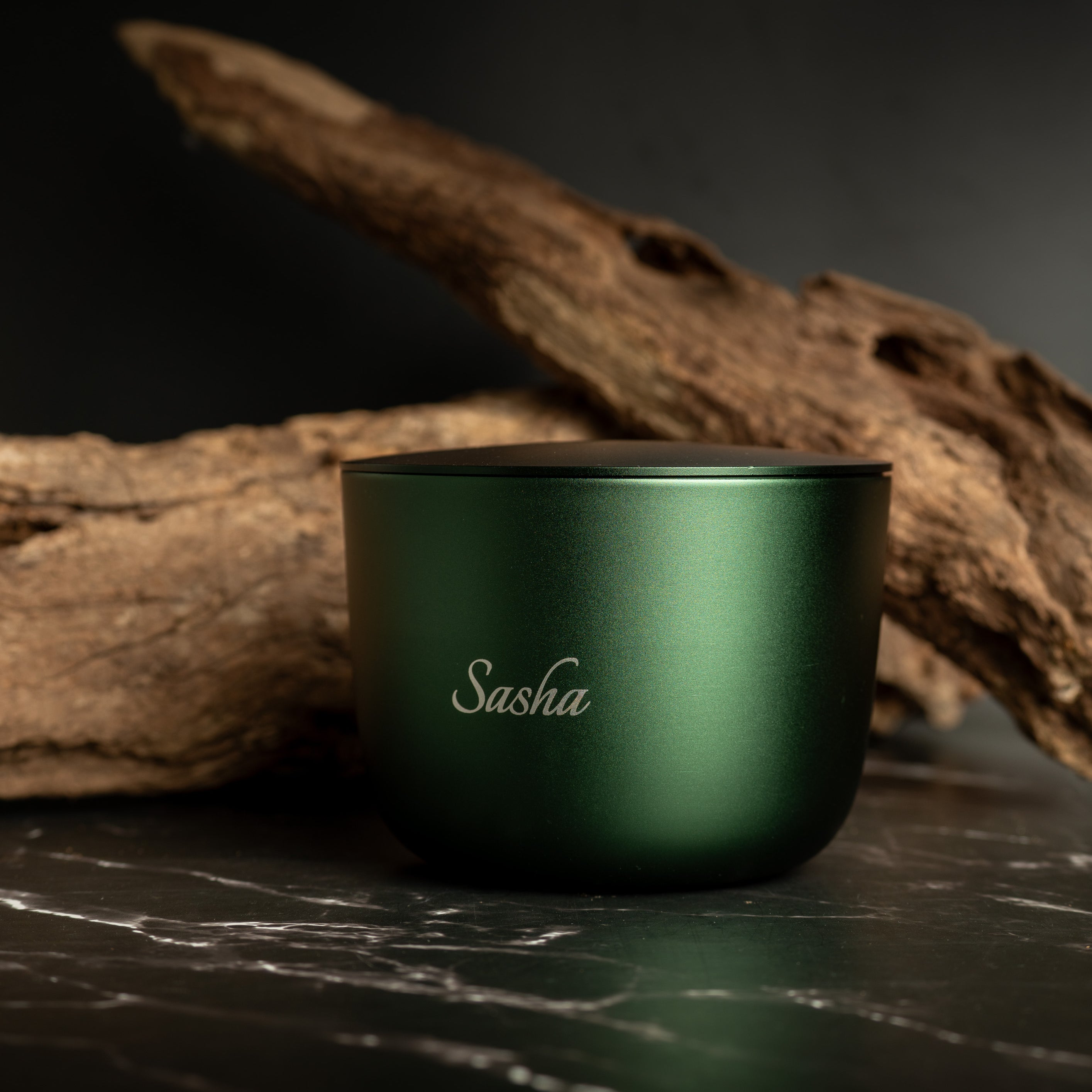 Green aluminium candle with 'Sasha' branding on a dark surface with wooden elements.