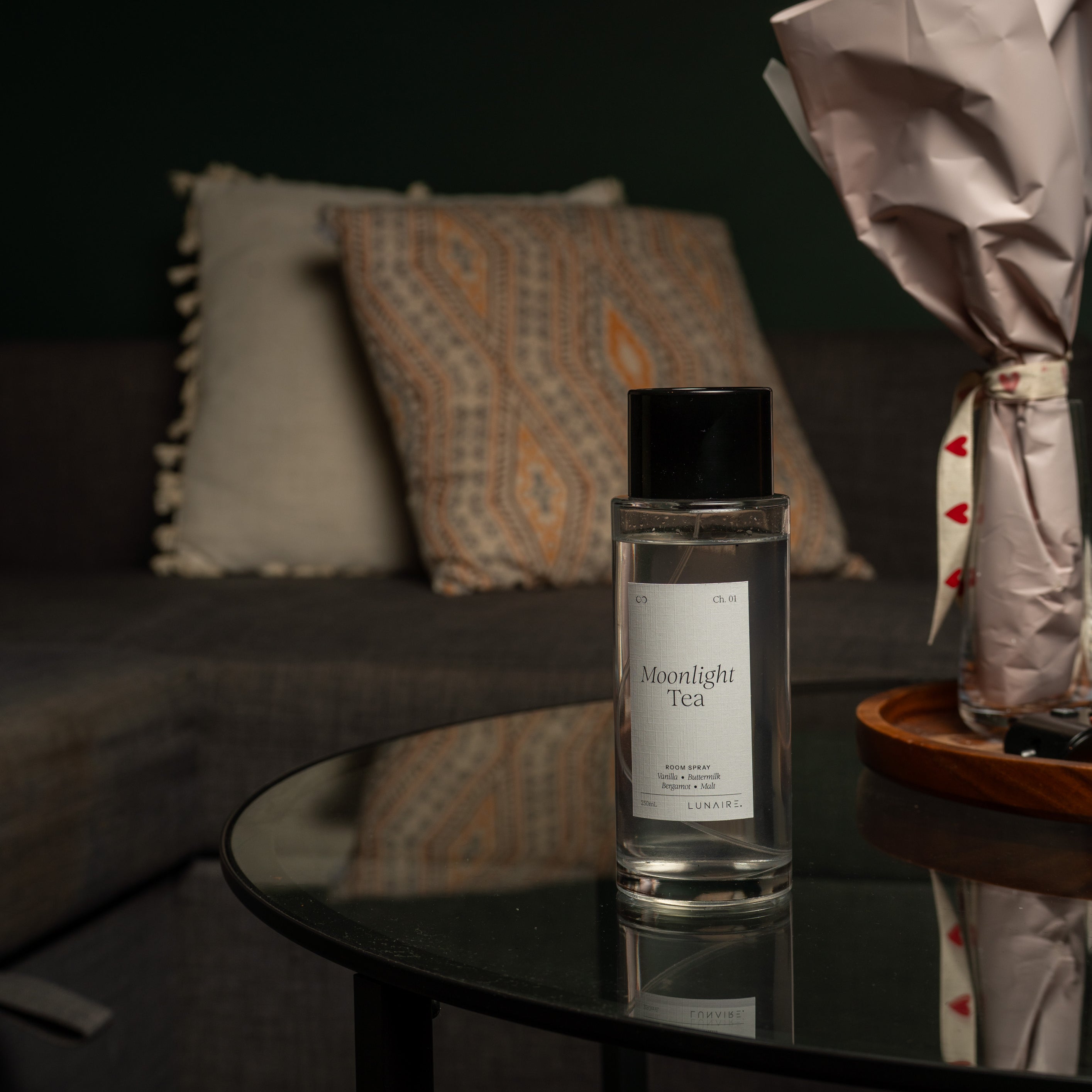 A clear glass bottle of Moonlight Tea room fragrance with a black cap and white label, placed on a round black glass table in a dimly lit living room setting.