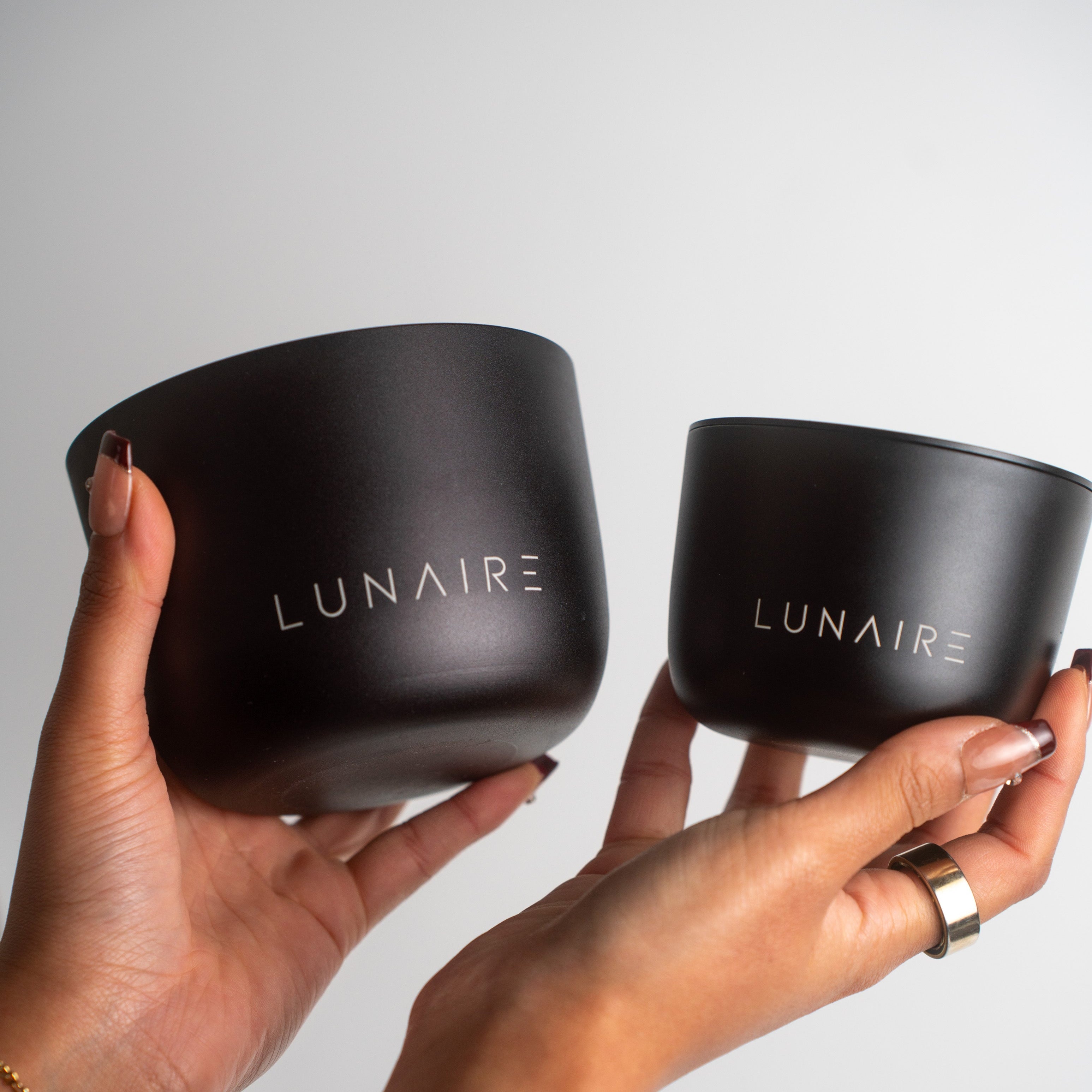 Two black aluminium luxury candle jars with 'LUNAIRE' branding held by two hands against a gray background.