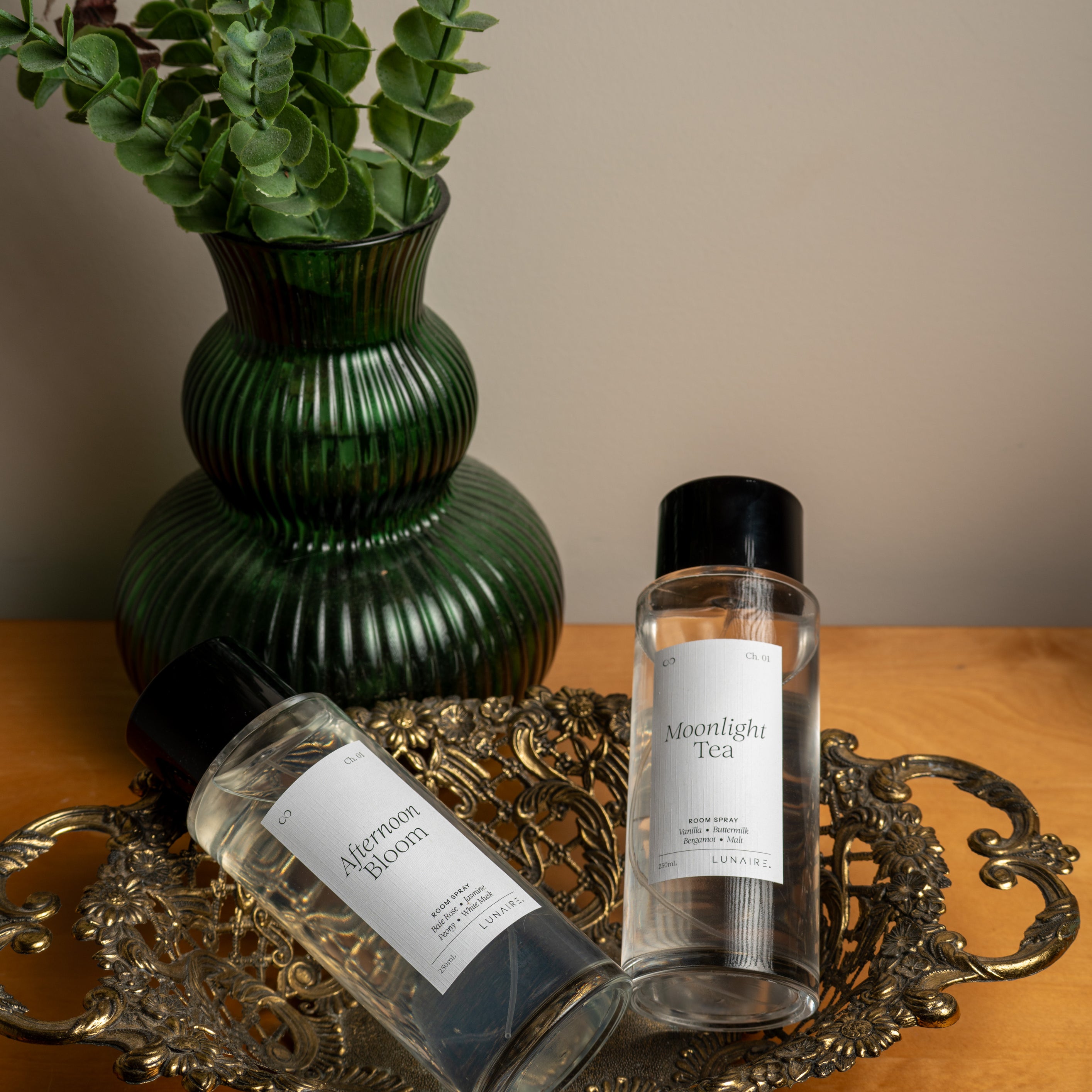 Two clear bottles with white labels and black caps, one reading 'Moonlight Tea' and the other 'Afternoon Bloom,' arranged on an ornate silver tray beside a green ribbed vase with leafy stems on a wooden surface. Room Refresher Spray by Lunaire