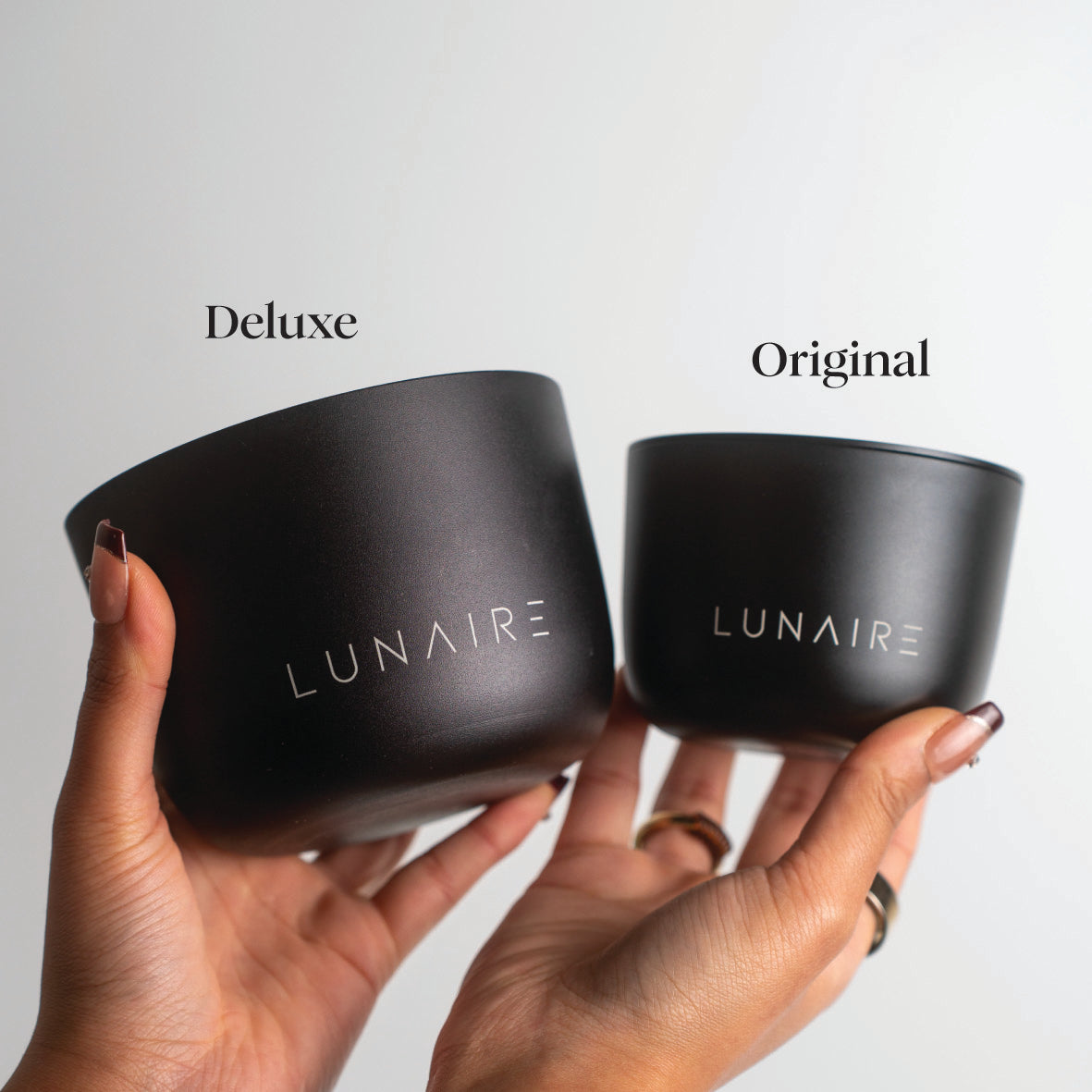 Two black aluminium candles labeled 'Deluxe' and 'Original' held by a hand against a light gray background.