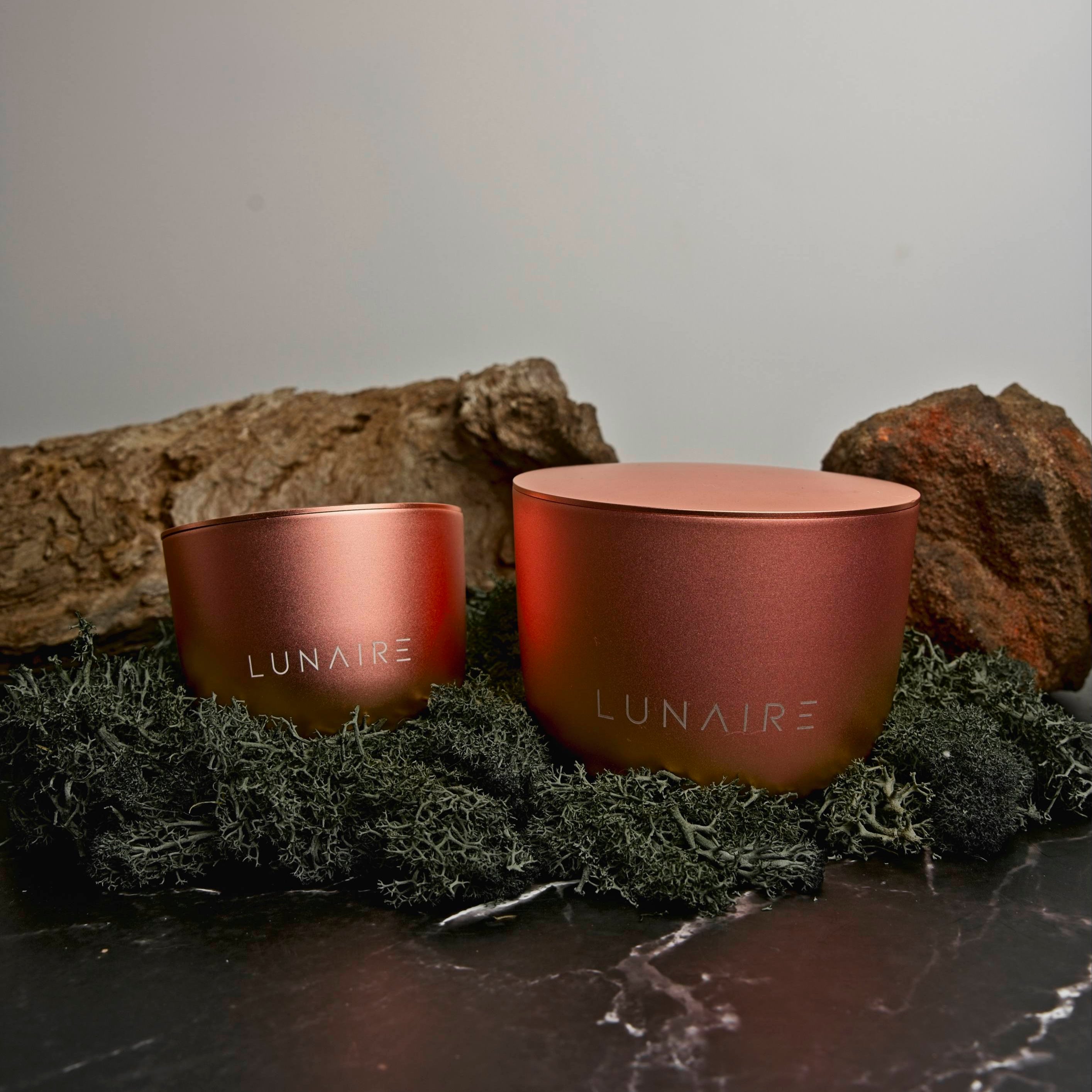Two rose gold-colored 'LUNAIRE' candles on a stone surface with moss and rocks in the background.