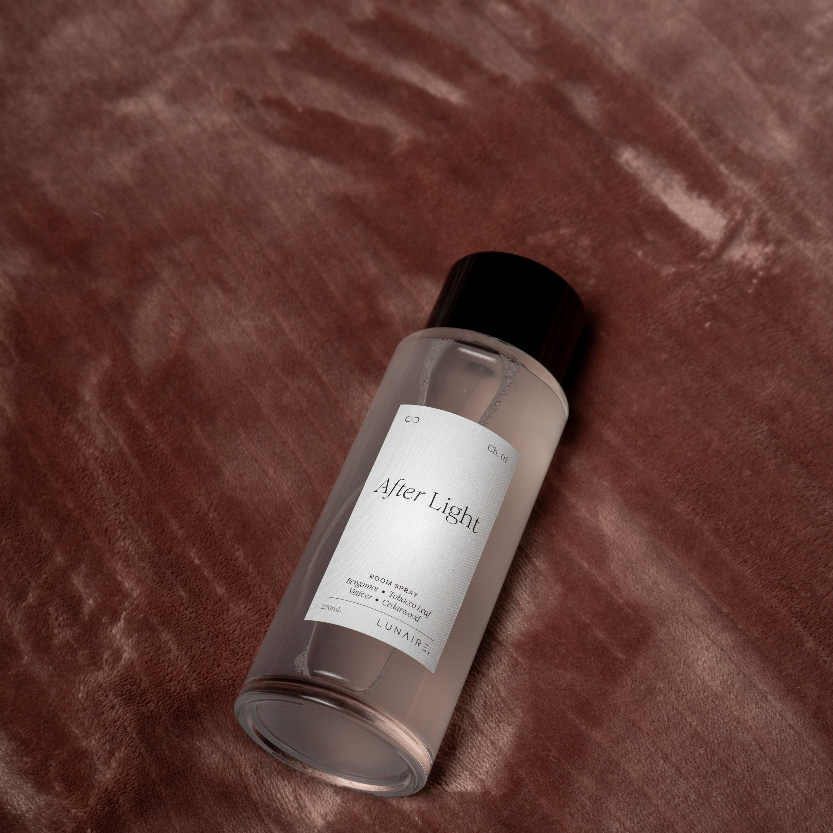 A clear cylindrical bottle with a black cap and a white label reading 'After Light' lies on a textured, dark brown velvet-like fabric background. linen Room Refresher Spray