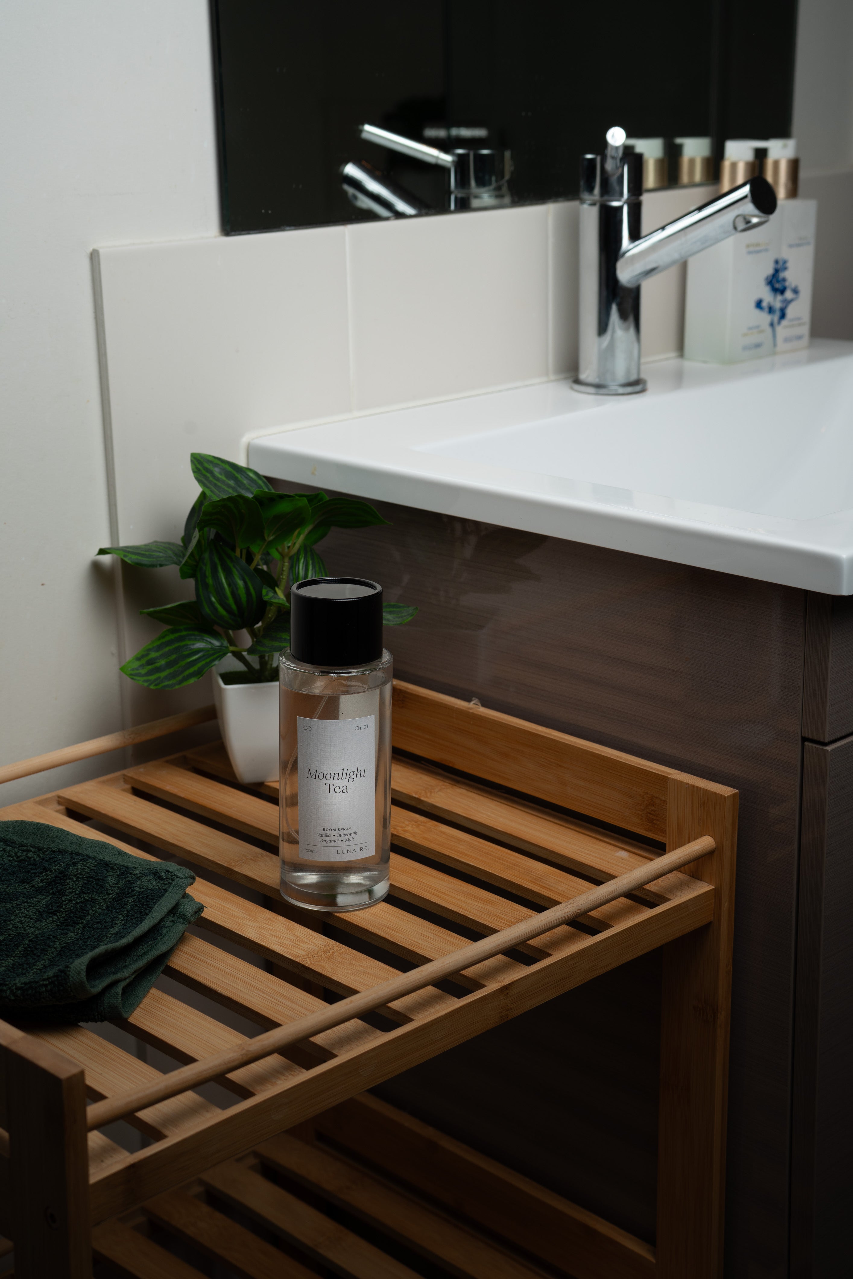 A clear glass bottle with a black cap and white label sits on a wooden slatted shelf beside a white sink, with a folded dark green towel and a small potted plant nearby. Room Refresher Spray