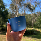 Hand holding a blue mug with 'Sarah & Oli' engraving against a natural background
