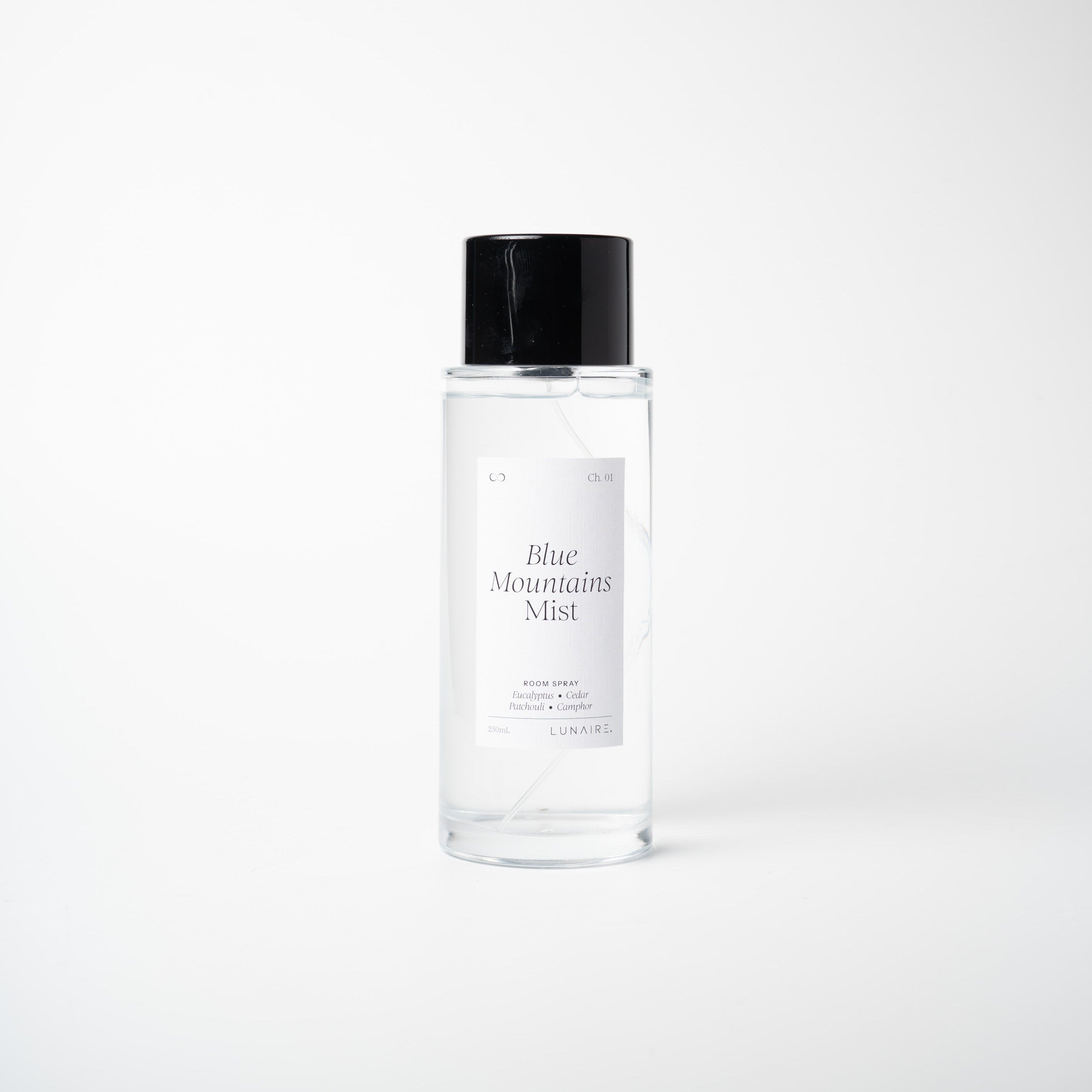 A clear glass bottle of Blue Mountain Mist room spray with a black cap and a white label, centered against a light gray background.
