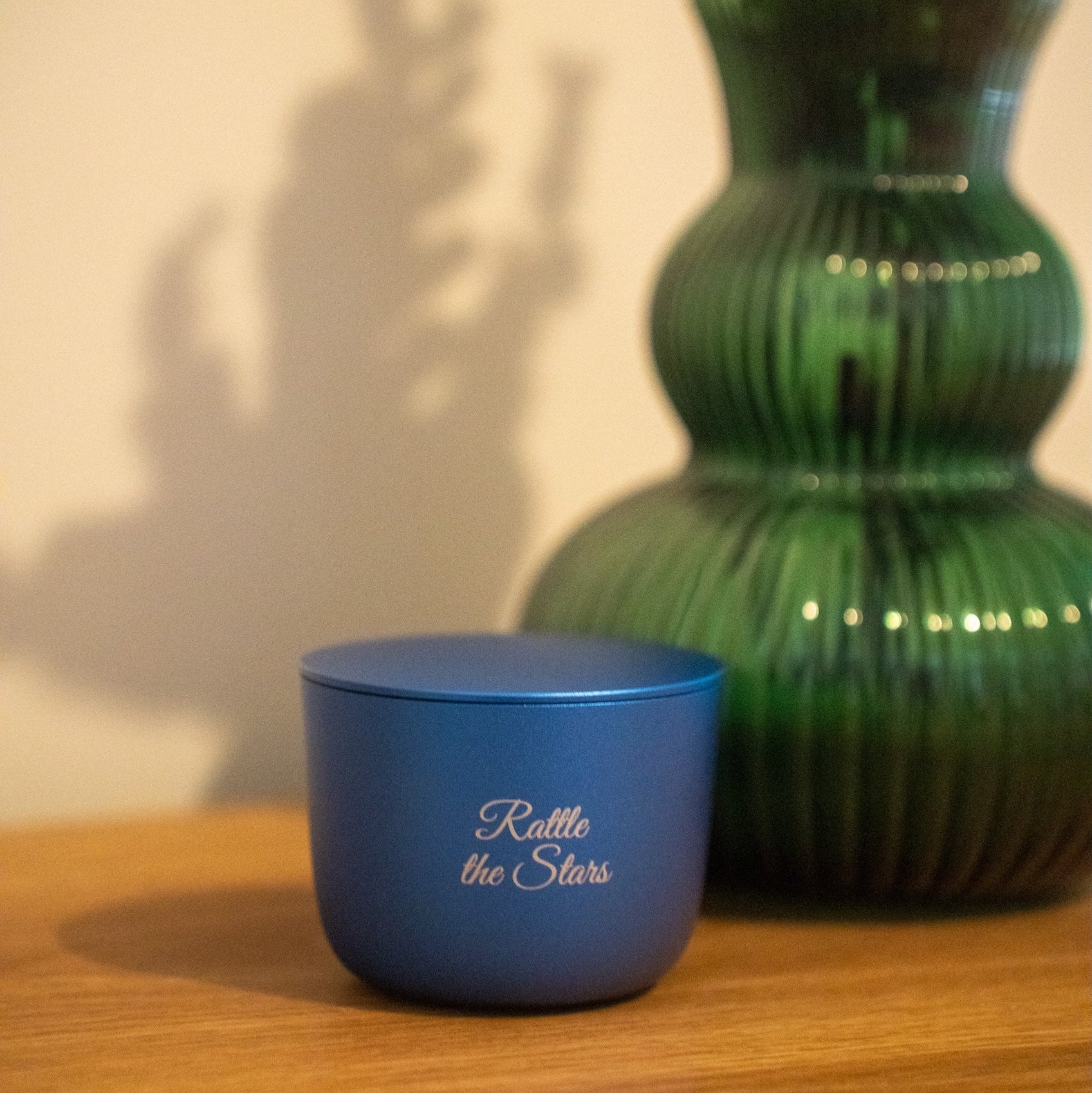 Blue candle vessel with 'Rattle the Stars' text next to a green vase on a wooden surface.