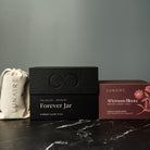 Lunaire aluminium candles including a black box labeled 'Forever Jar' and a red box labeled 'Afternoon Bloom' on a dark surface. luxury gift. 
