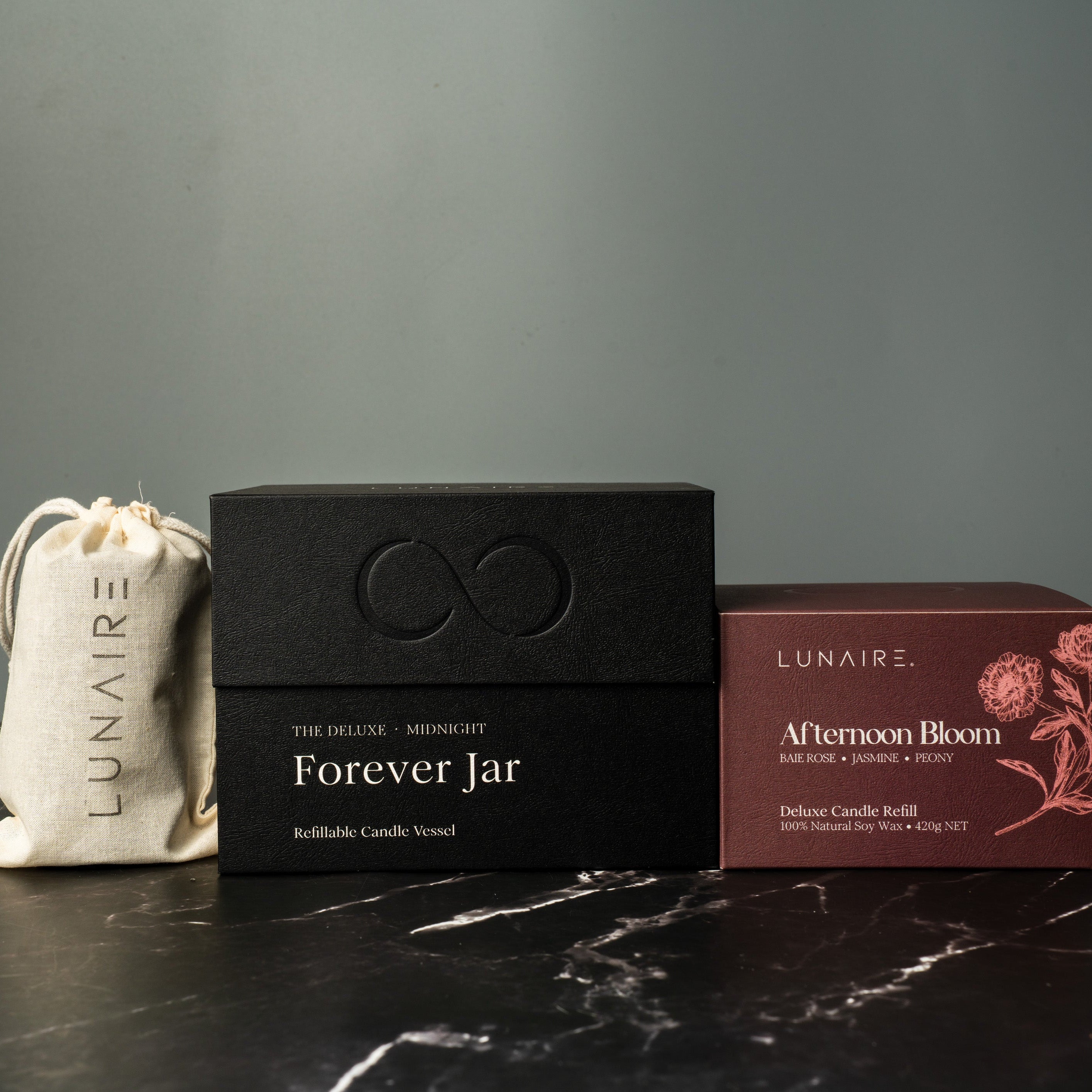 Lunaire aluminium candles including a black box labeled 'Forever Jar' and a red box labeled 'Afternoon Bloom' on a dark surface. luxury gift. 