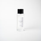 A clear glass bottle of 'First Light Fern' room spray with a black cap and a white label, set against a seamless light gray background.