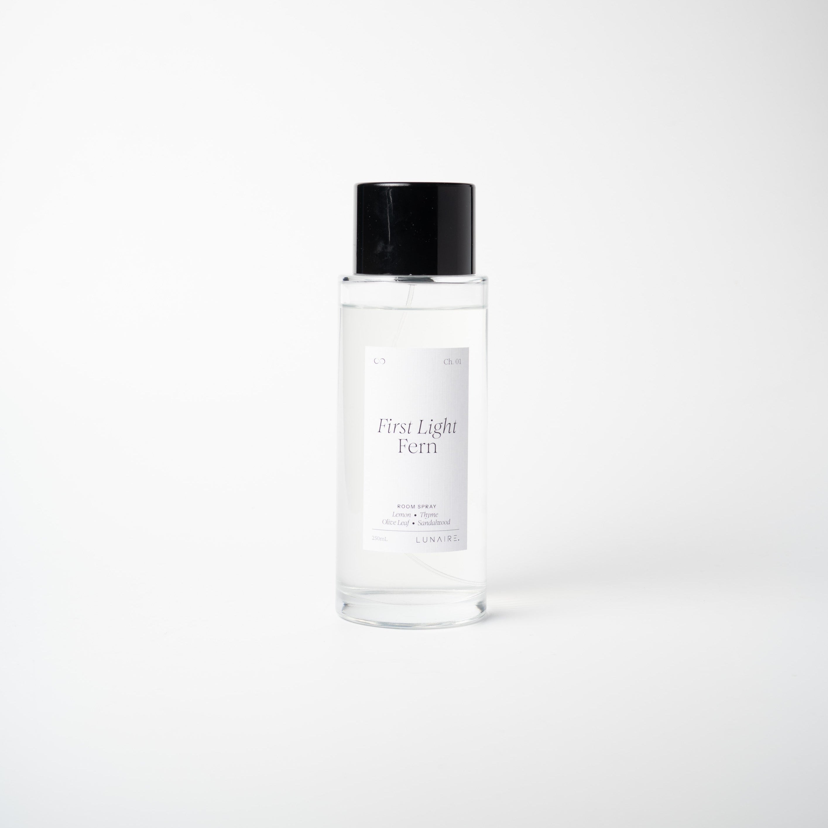 A clear glass bottle of 'First Light Fern' room spray with a black cap and a white label, set against a seamless light gray background.
