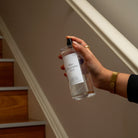A hand holding a clear bottle of Lunaire Room Refresher Spray against an indoor staircase background.