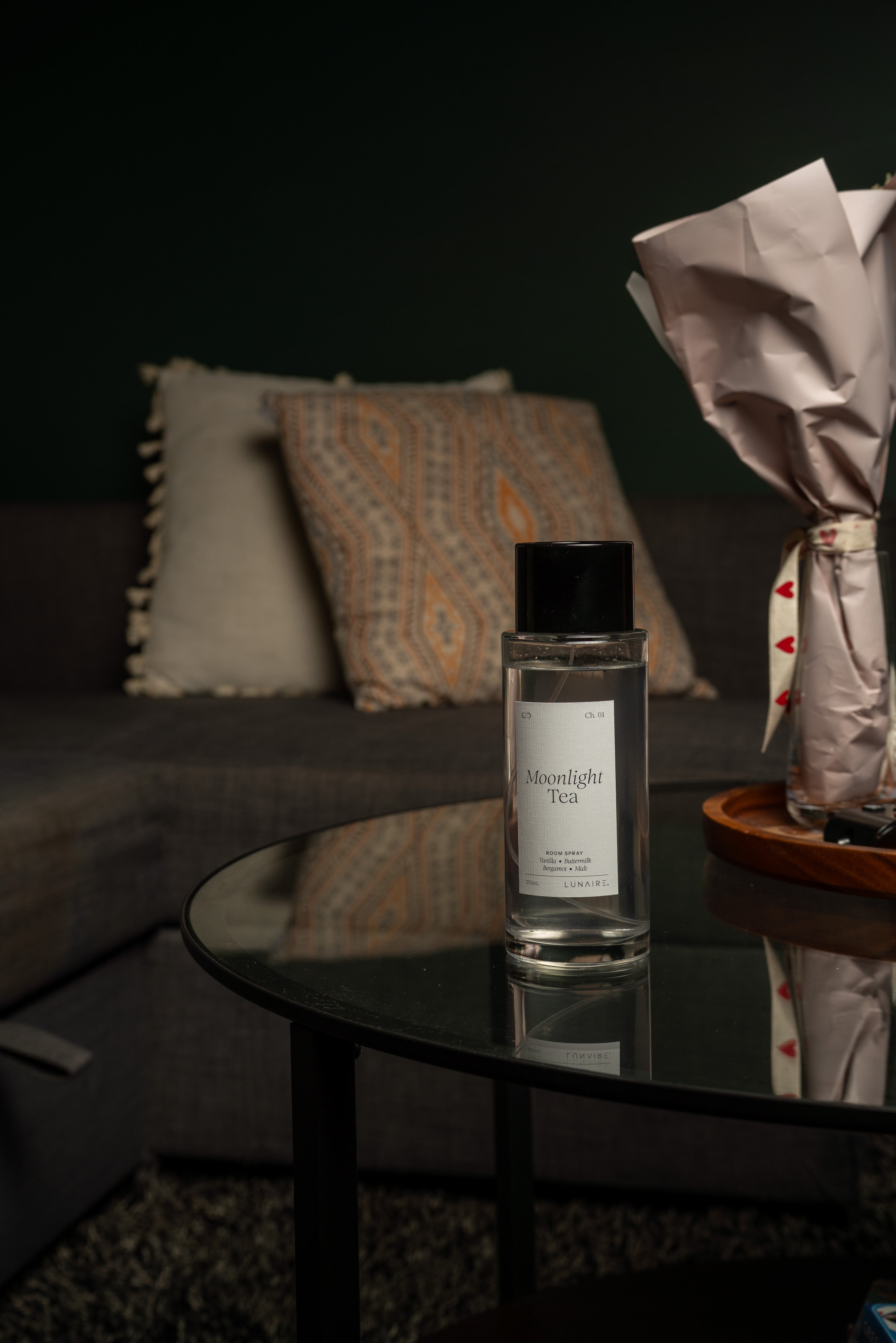 A clear glass bottle of Moonlight Tea room fragrance with a black cap and white label, placed on a round black glass table in a dimly lit living room setting.