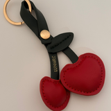 Red cherry-shaped keychain with green strap and gold ring on a light gray background