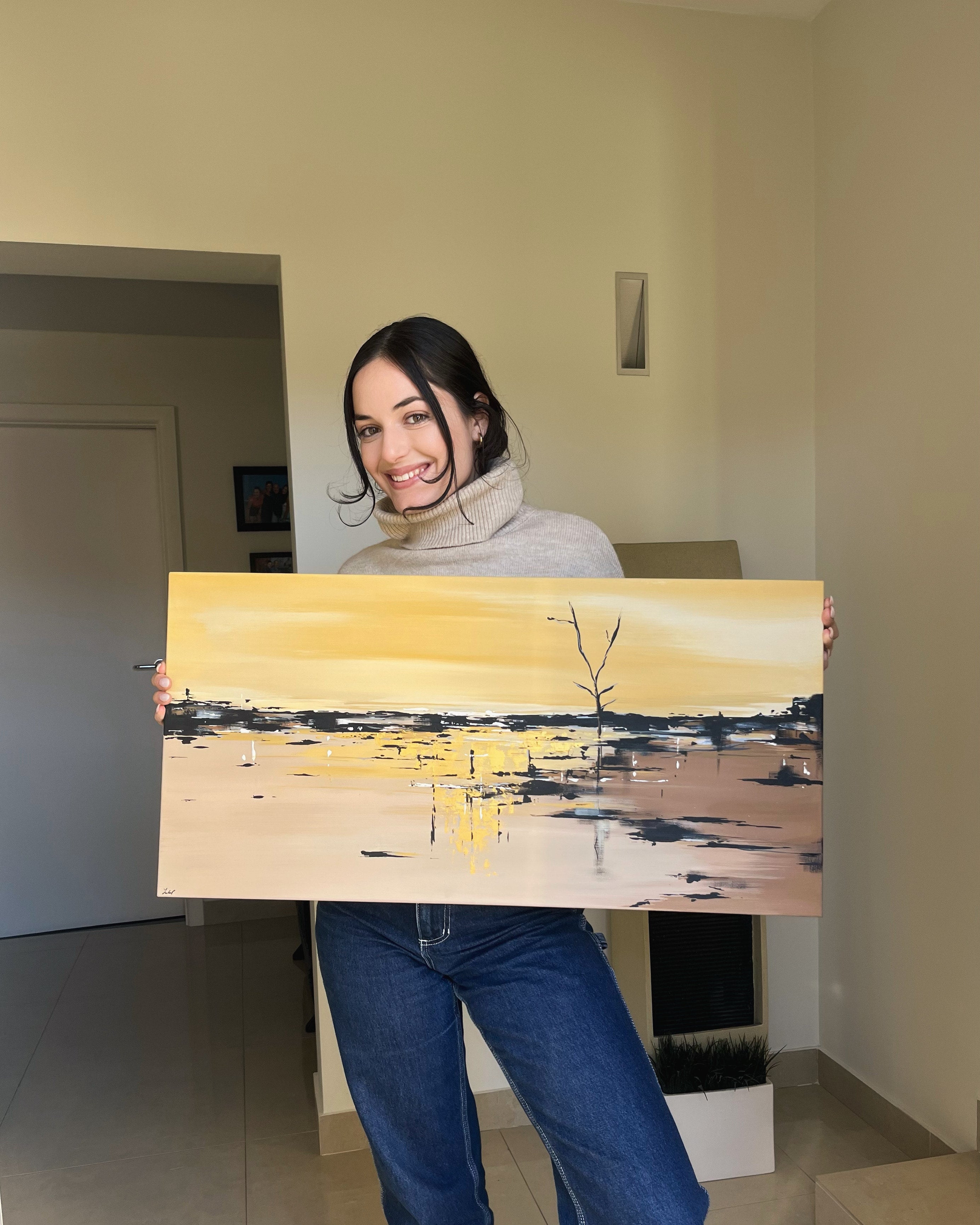 Artist Julia Parisi holding a painting with a yellow landscape scene indoors.