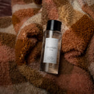 A clear bottle of Moonlight Tea room spray with a black cap and white label, resting on a cozy, textured striped blanket in warm brown, beige, and rust tones.