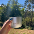 Hand holding a silver Lunaire Silver Aluminium candle with 'MUM' engraved, outdoors with trees in the background