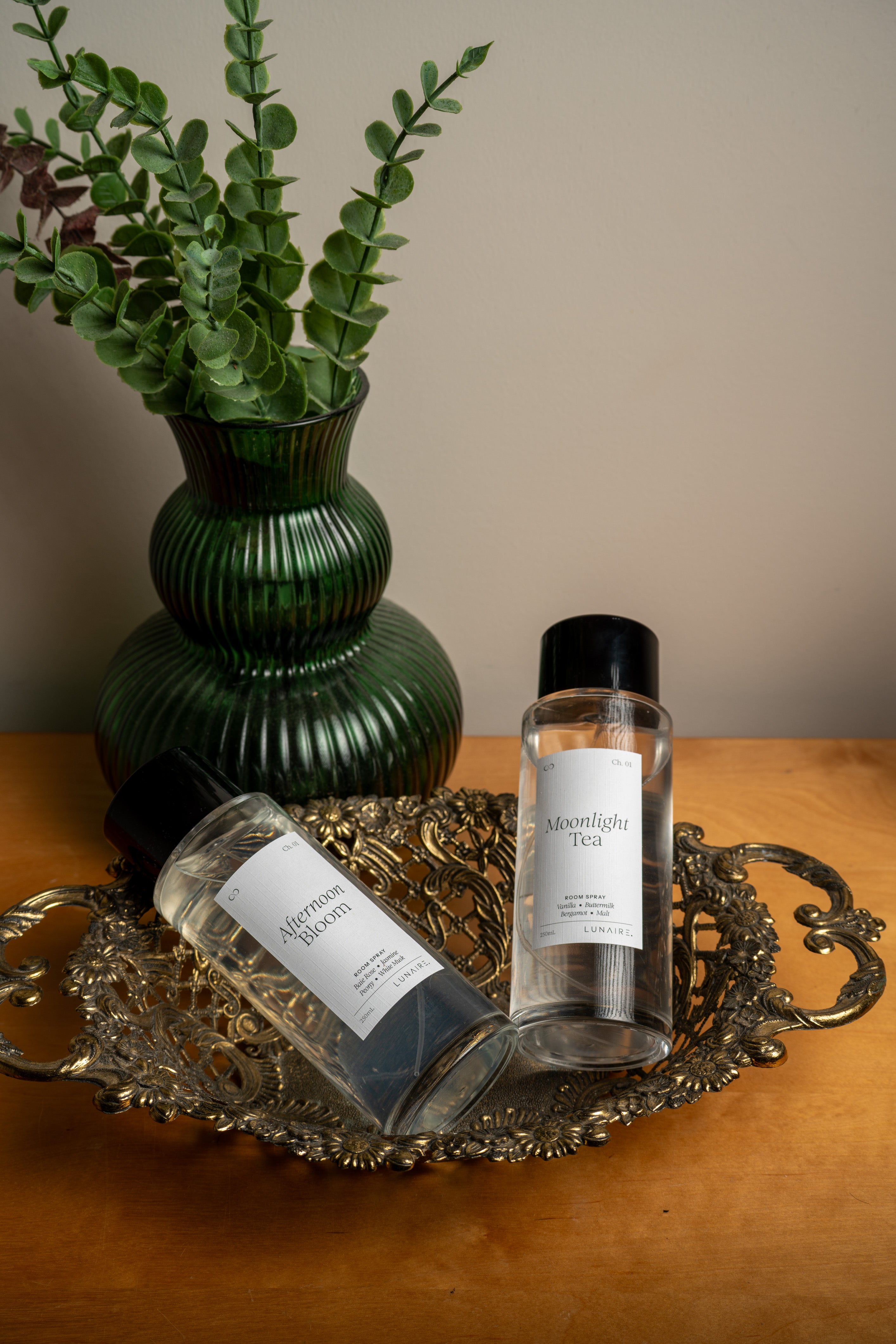 Two clear bottles with white labels and black caps, one reading 'Moonlight Tea' and the other 'Afternoon Bloom,' arranged on an ornate silver tray beside a green ribbed vase with leafy stems on a wooden surface. Room Refresher Spray by Lunaire