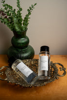 Two clear bottles with white labels and black caps, one reading 'Moonlight Tea' and the other 'Afternoon Bloom,' arranged on an ornate silver tray beside a green ribbed vase with leafy stems on a wooden surface. Room Refresher Spray by Lunaire