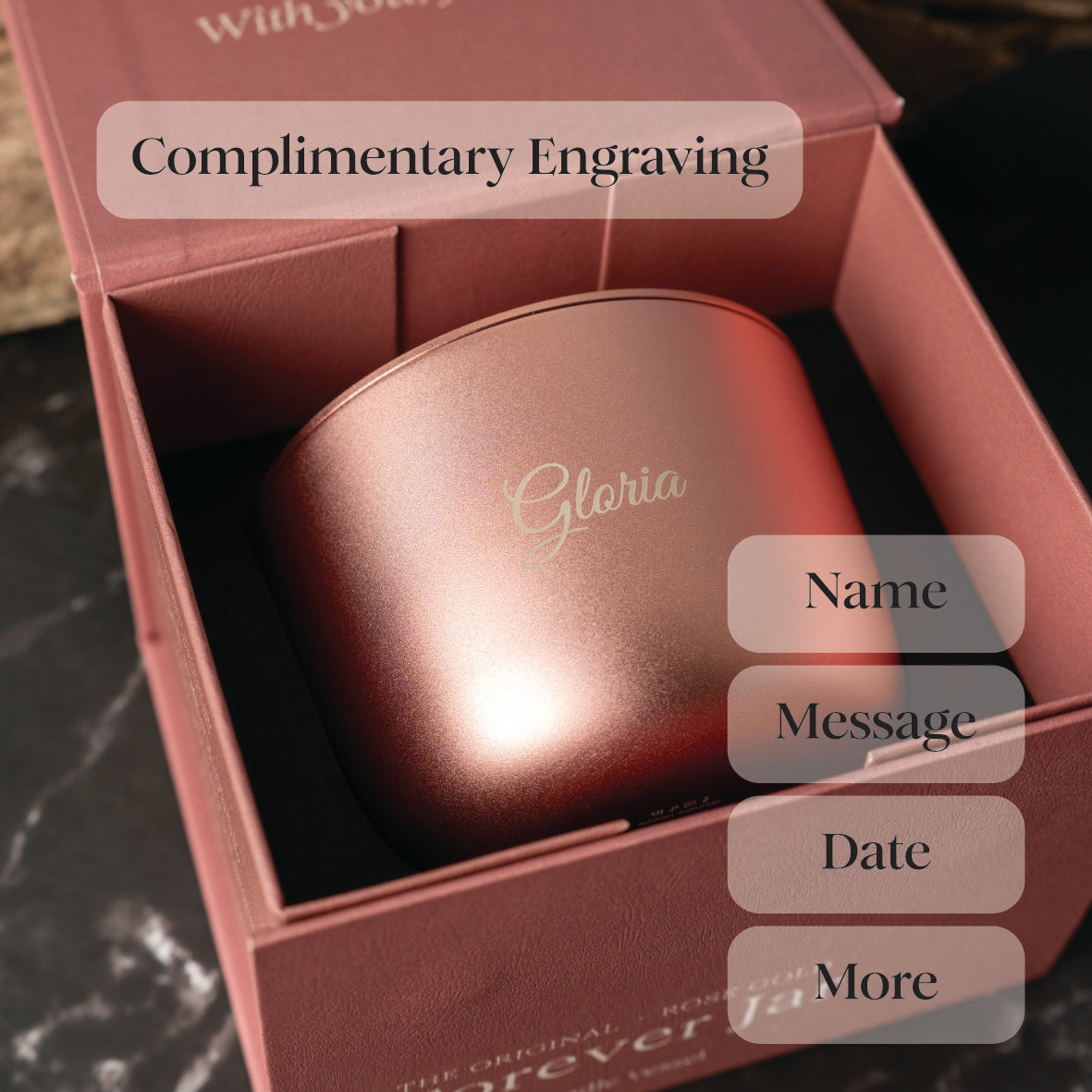 Rose gold luxury candle with 'Gloria' engraving in a pink box, on a dark surface.