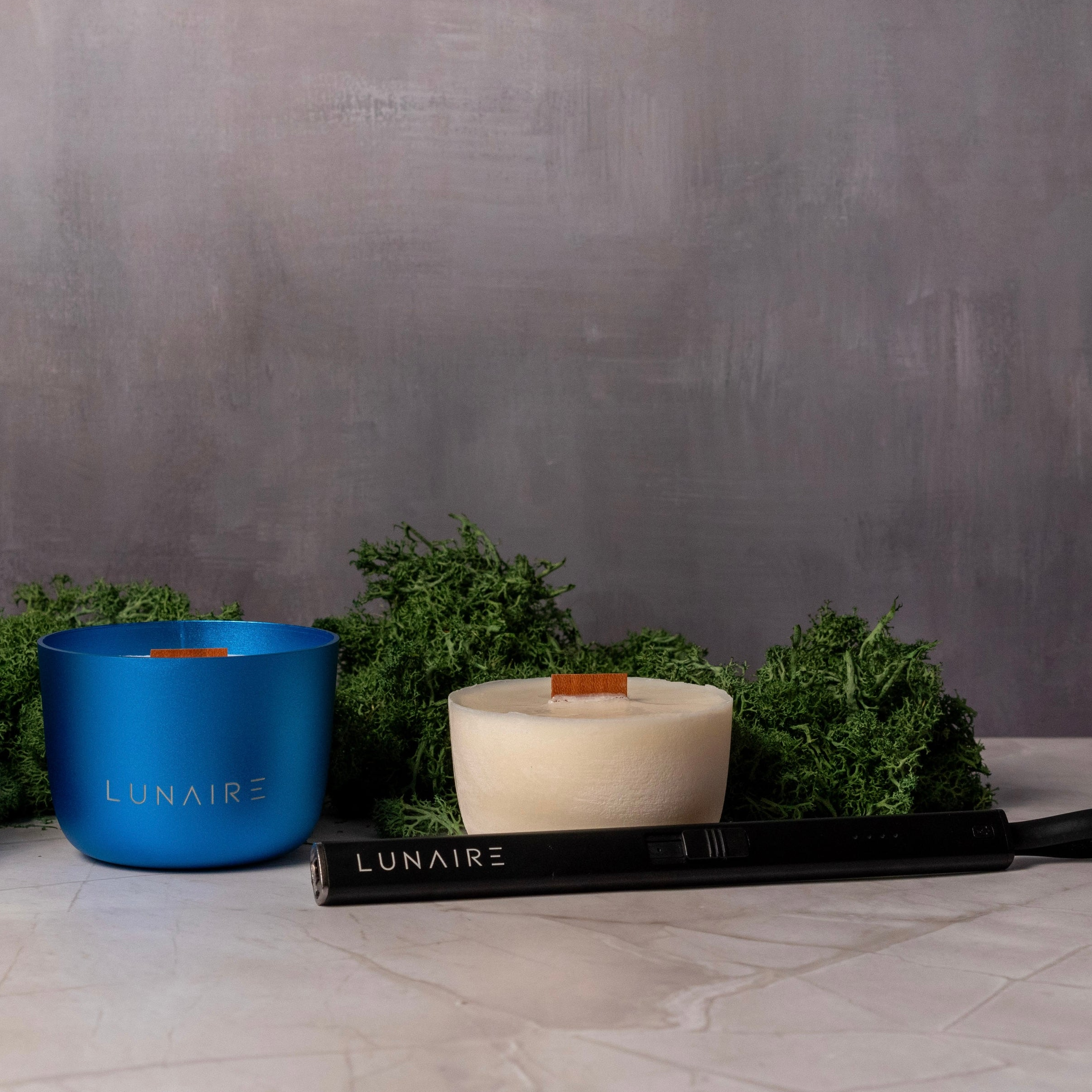 Blue refillable Forever Candle with wood wick next to refill and rechargeable arc lighter. Surrounded by greenery 