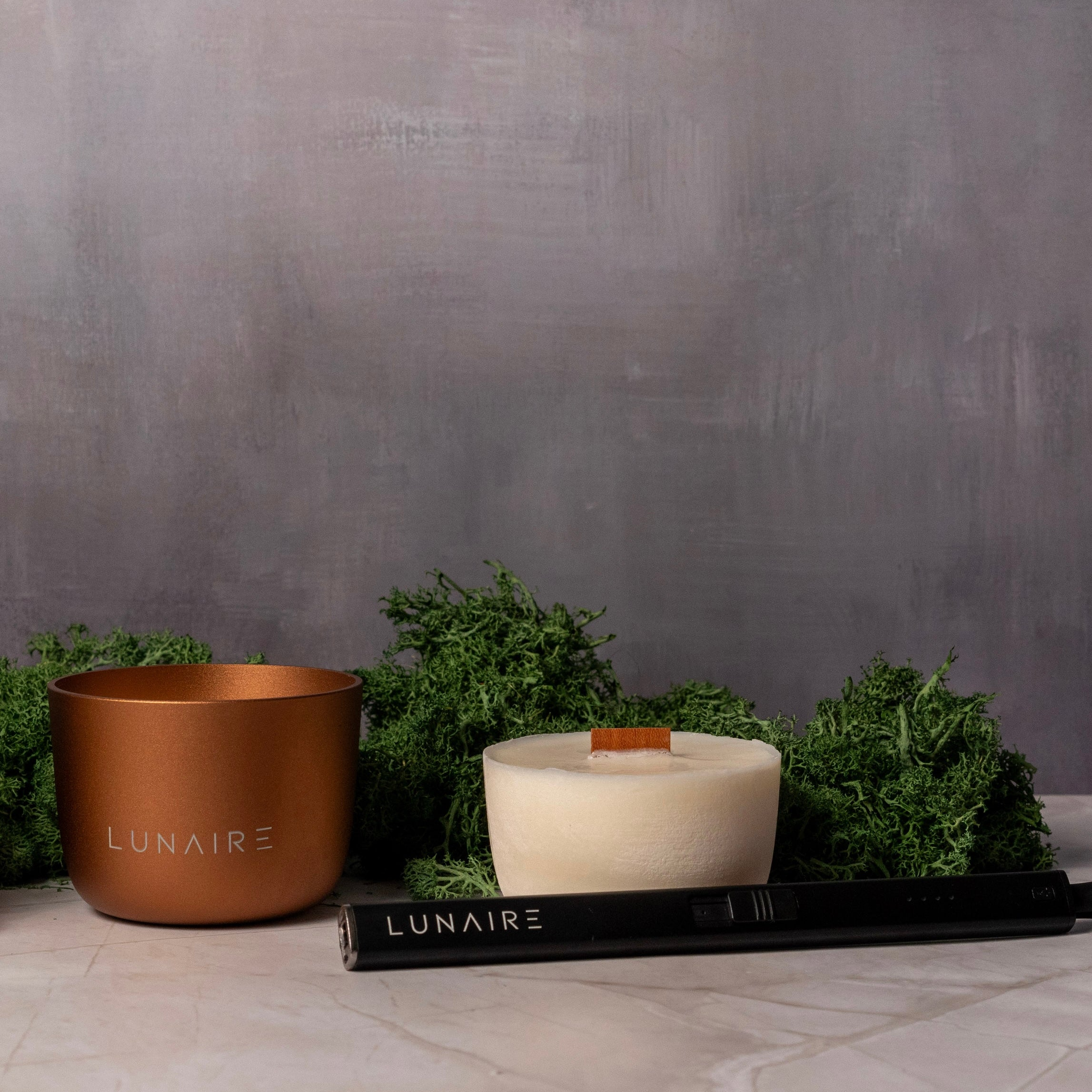 Bronze refillable Forever Candle with wood wick next to refill and rechargeable arc lighter. Surrounded by greenery 