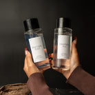 Two clear glass bottles with black caps and white labels are held up by two hands against a dark background. The left bottle is labeled 'First Light Fern' and the right bottle is labeled 'Moonlight Tea'.