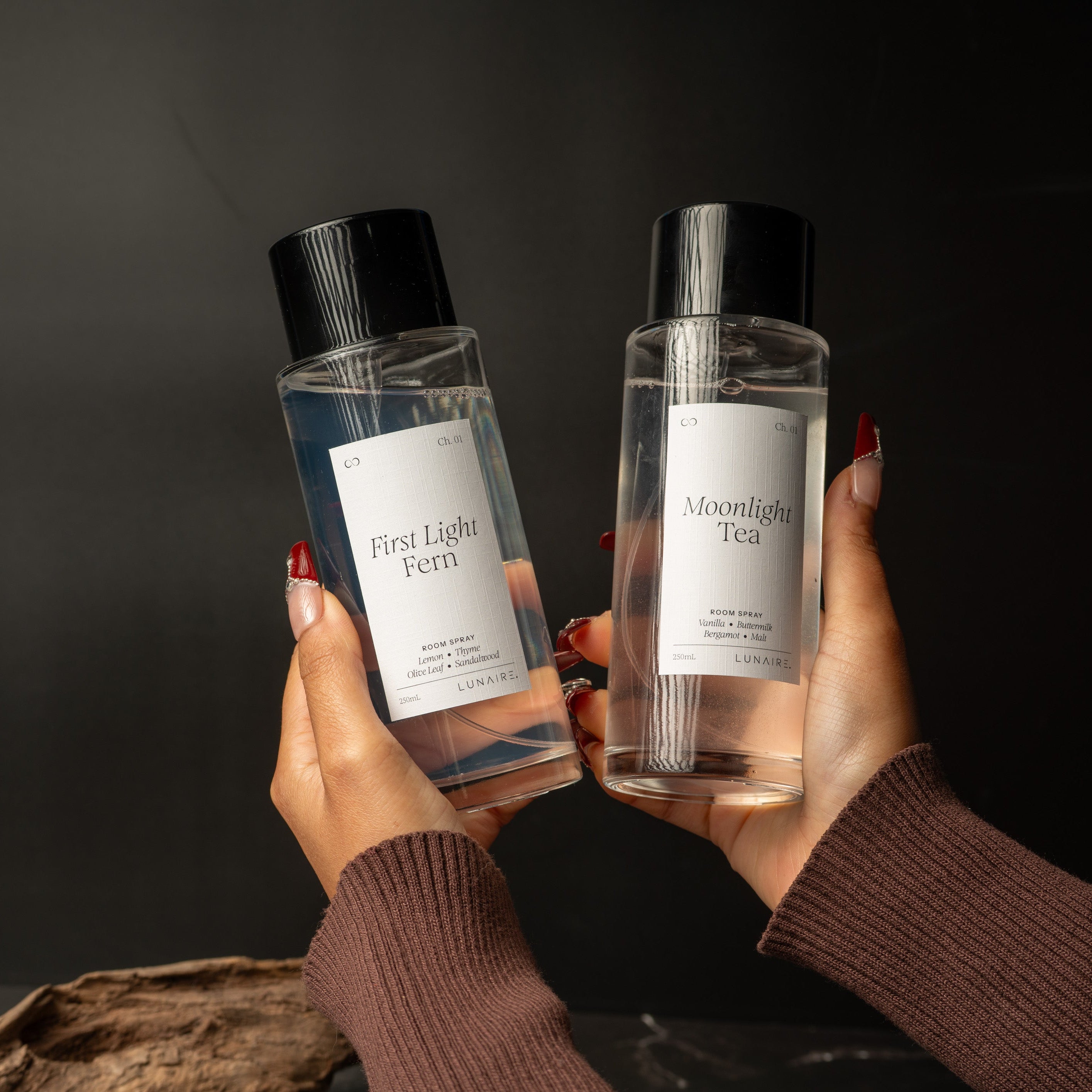 Two clear glass bottles with black caps and white labels are held up by two hands against a dark background. The left bottle is labeled 'First Light Fern' and the right bottle is labeled 'Moonlight Tea'.