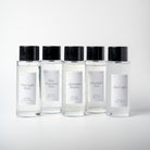 Five clear room spray bottles with black caps and white labels arranged in a row on a light gray background. Luxury home spray by Lunaire