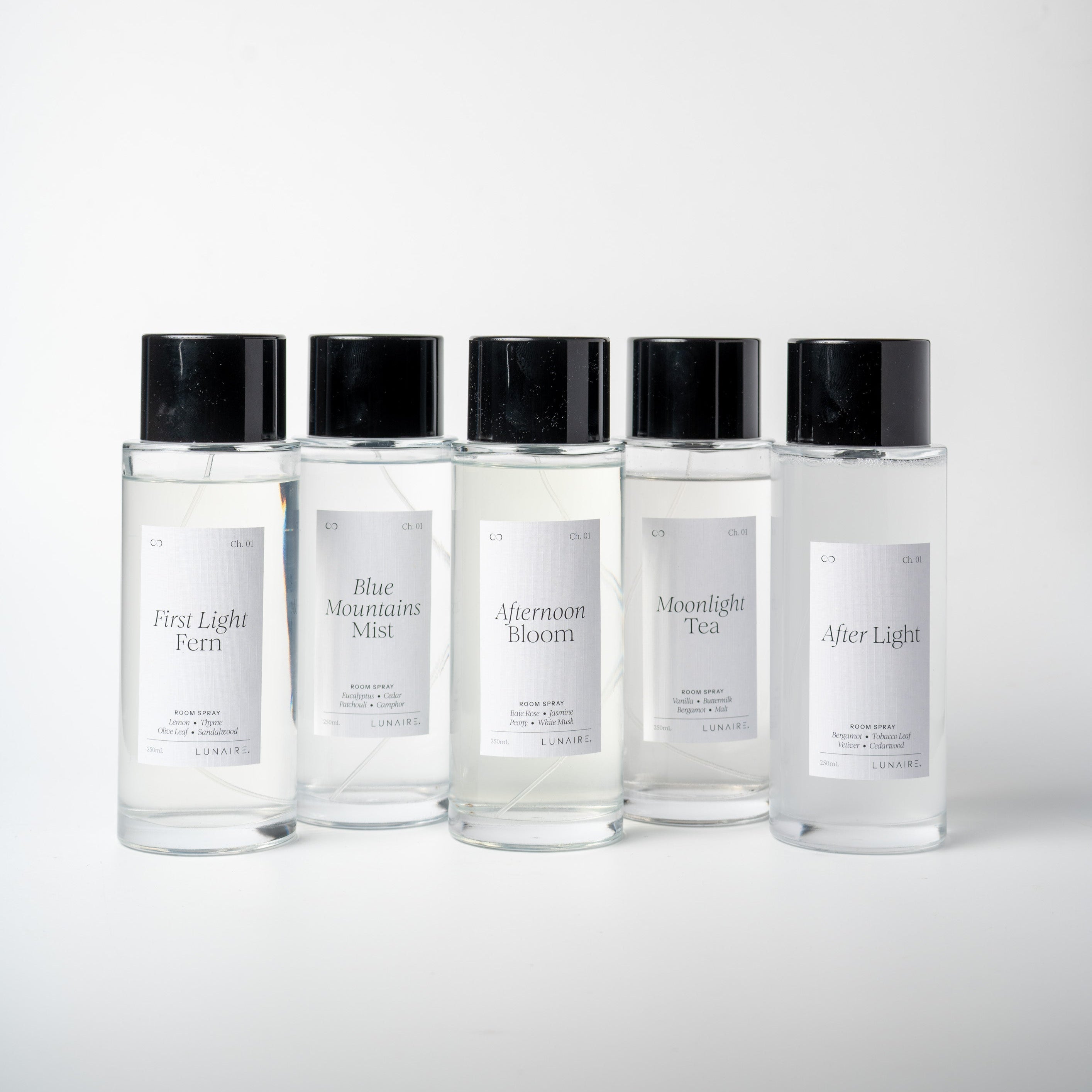 Five clear room spray bottles with black caps and white labels arranged in a row on a light gray background. Luxury home spray by Lunaire