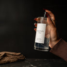 A hand with red manicured nails holds a clear glass spray bottle labeled 'Moonlight Tea' against a dark background, with a piece of driftwood on a dark surface in the foreground.