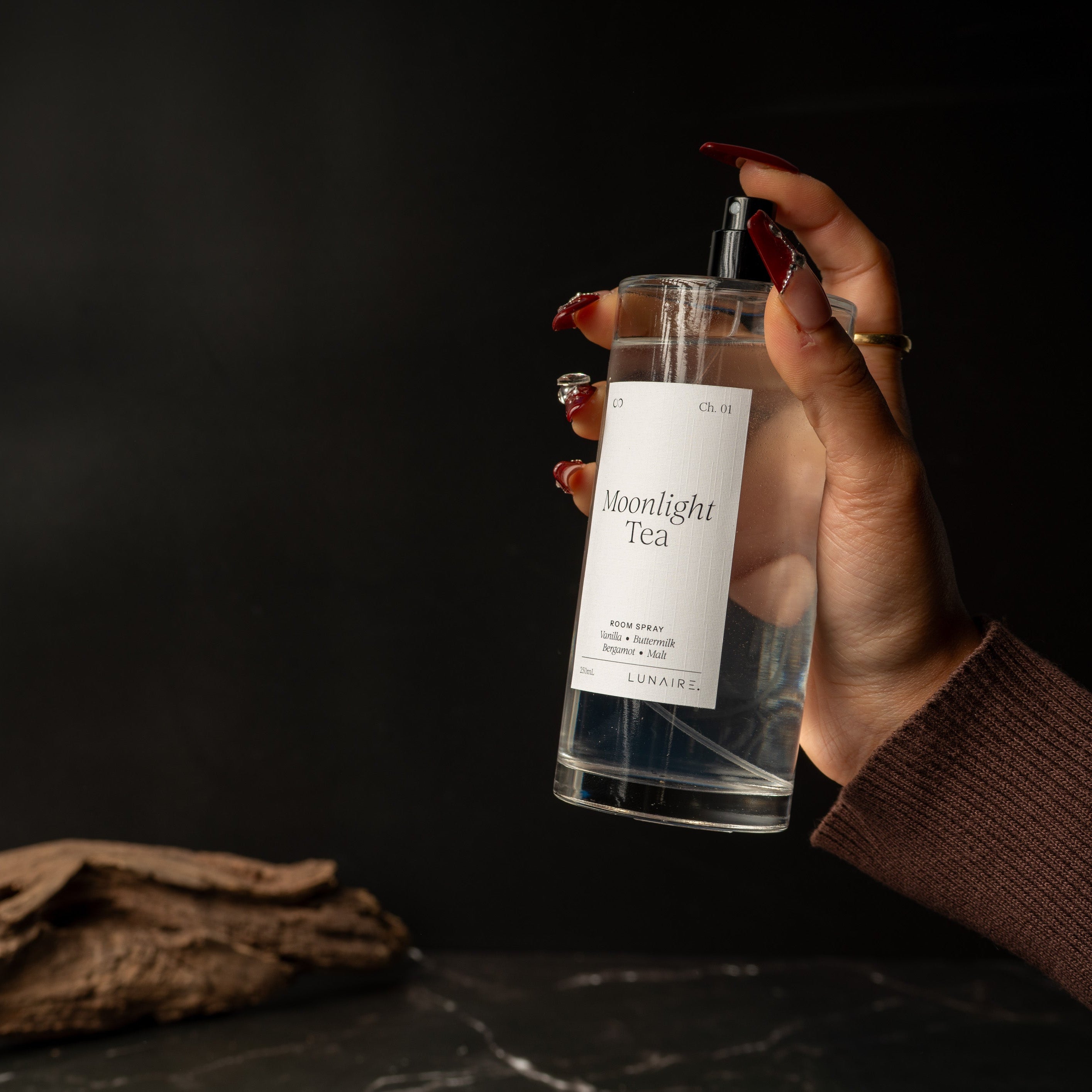 A hand with red manicured nails holds a clear glass spray bottle labeled 'Moonlight Tea' against a dark background, with a piece of driftwood on a dark surface in the foreground.