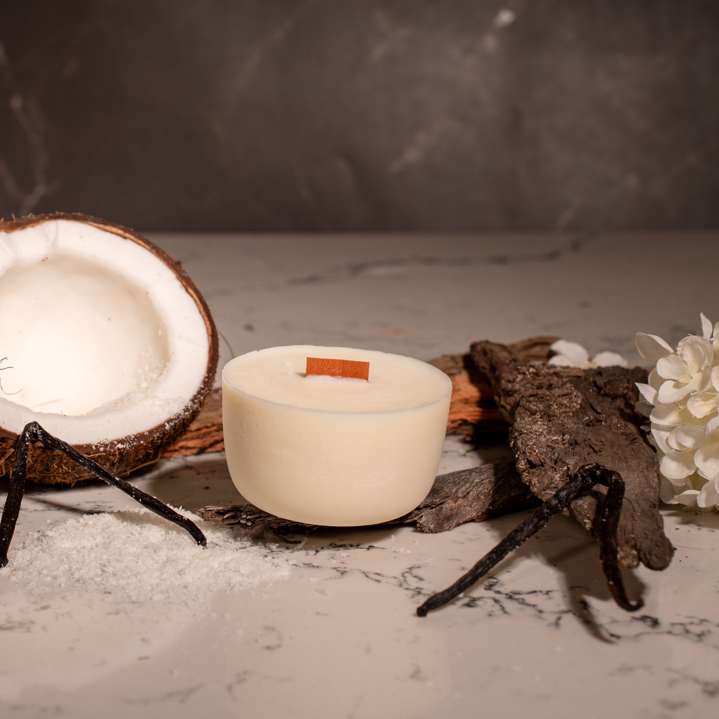 Coconut halves, a soy candle refill, and white flowers on a textured surface with a dark background.